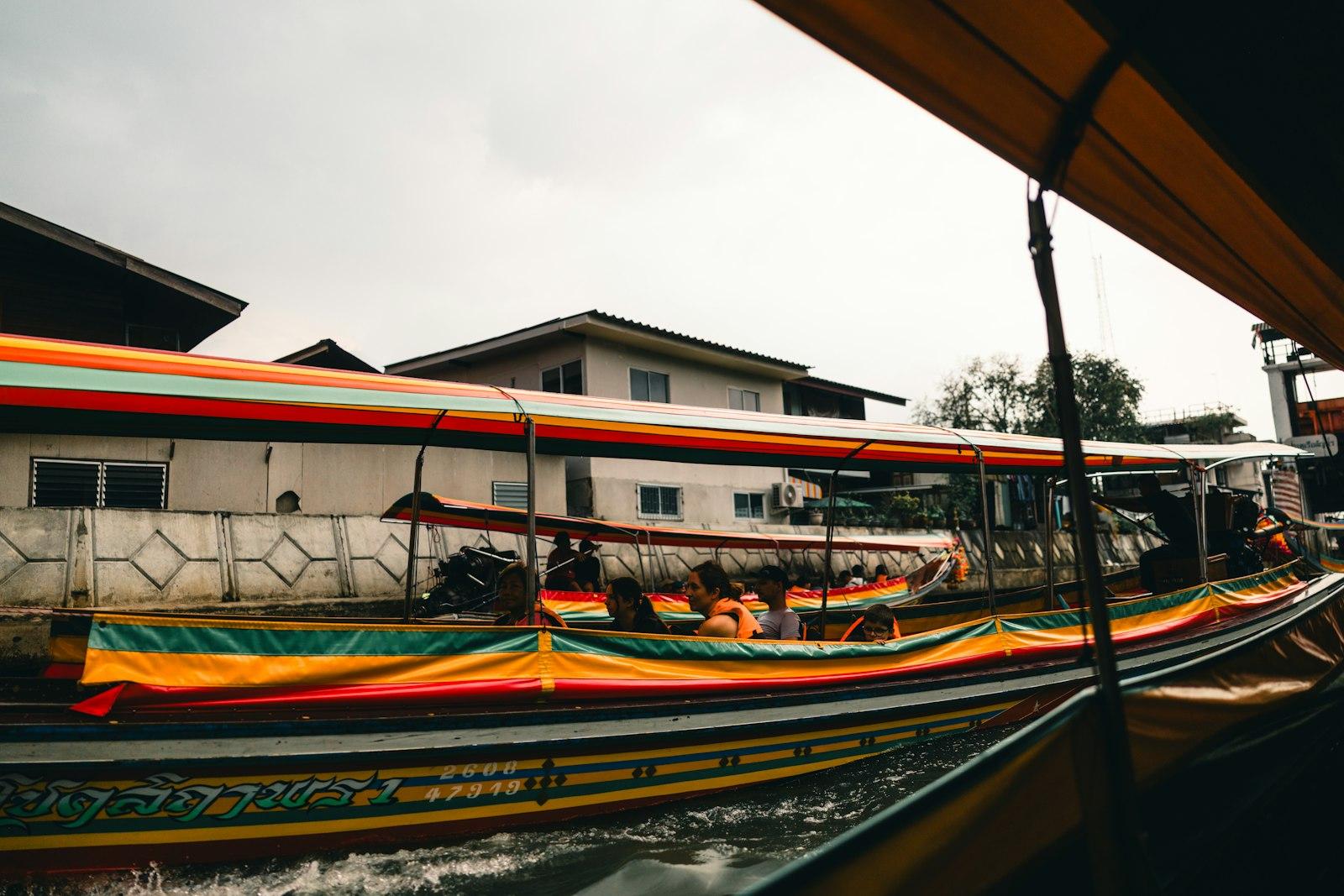 A brightly painted passenger longtail boat carrying people along a narrow Bangkok canal lined with houses