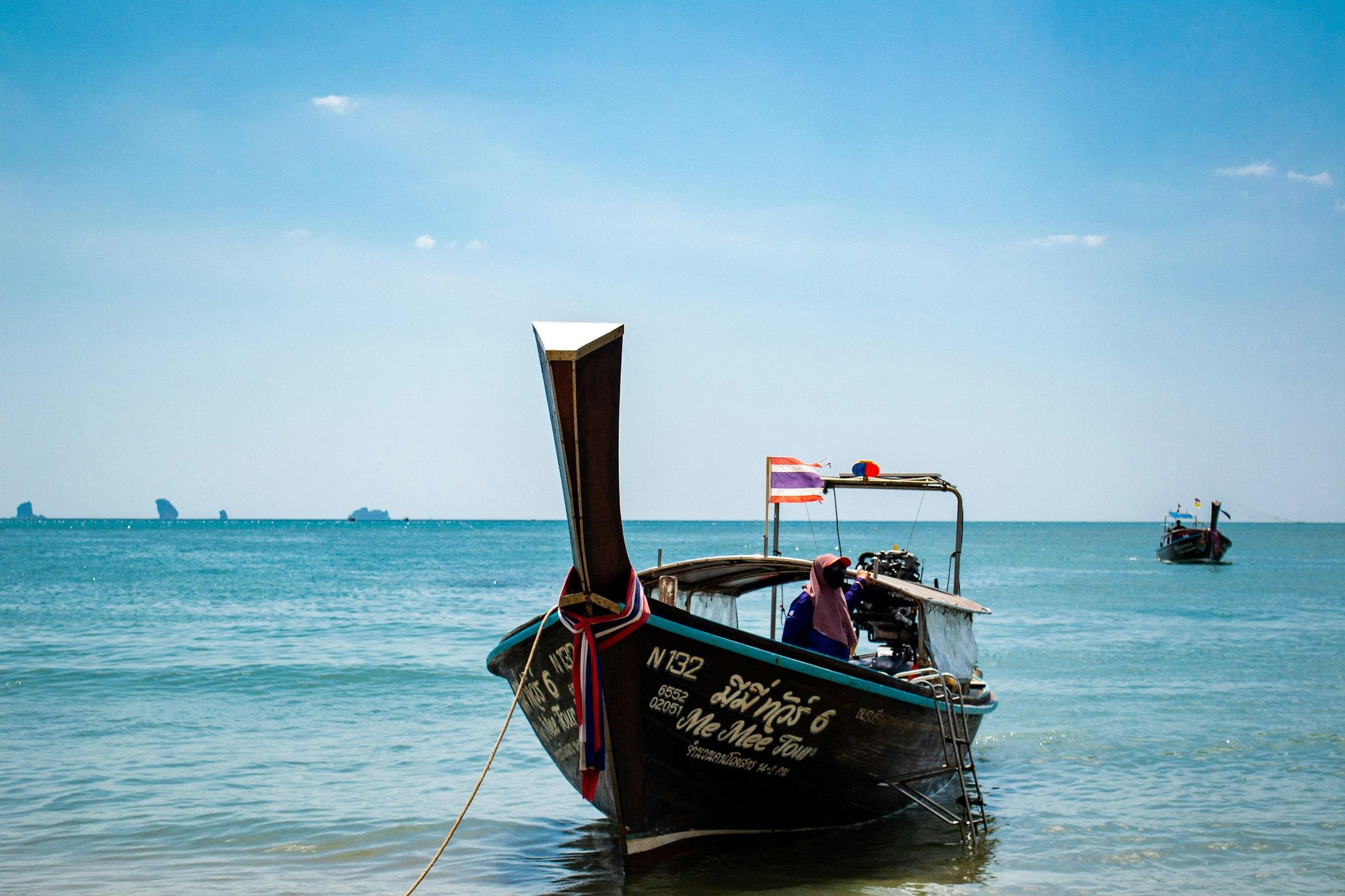 A wooden tour longtail boat at anchor in clear blue water, crew member aboard, karst islands on the horizon