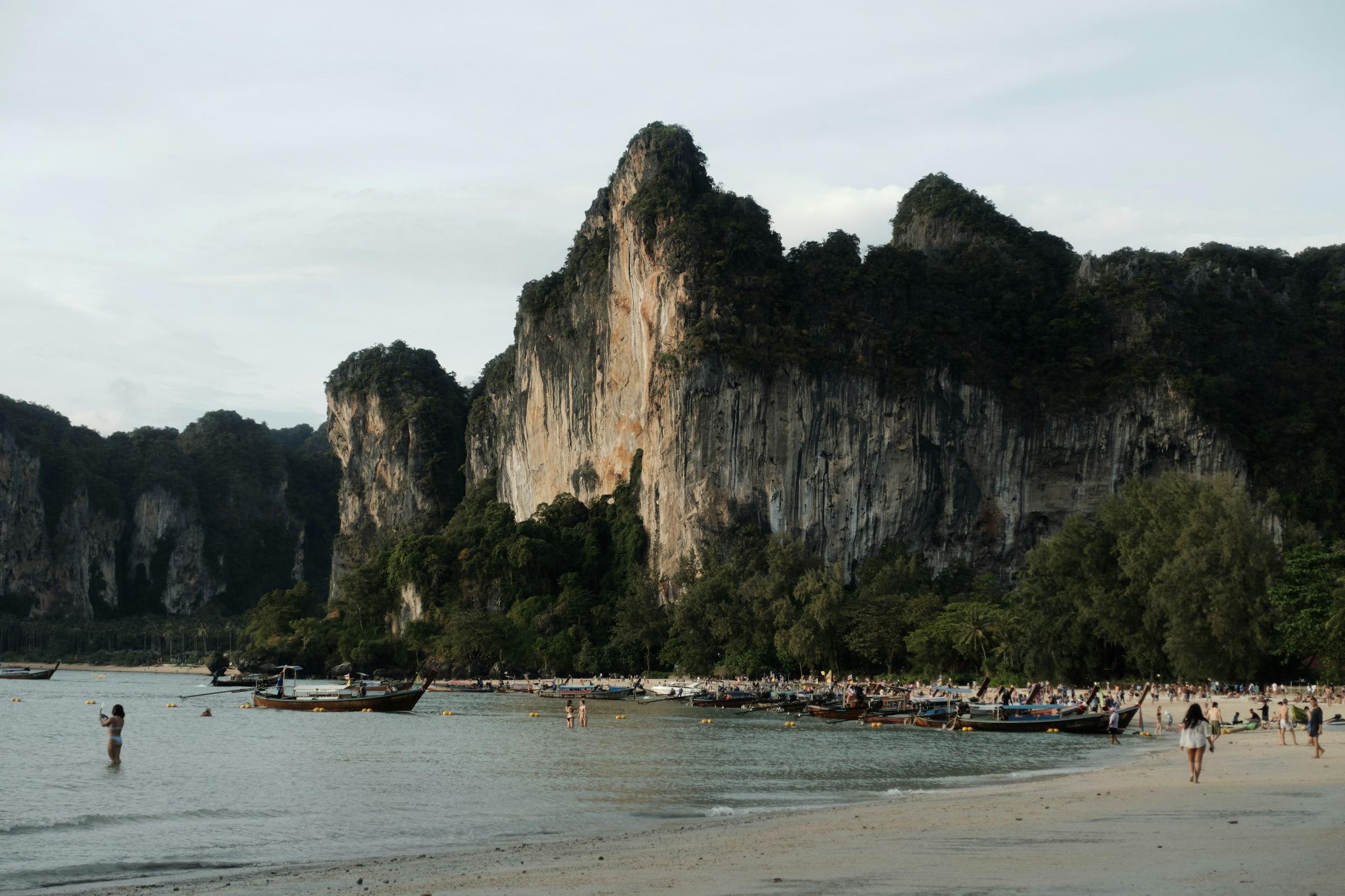 Longtail boats moored along a beach beneath a towering limestone cliff