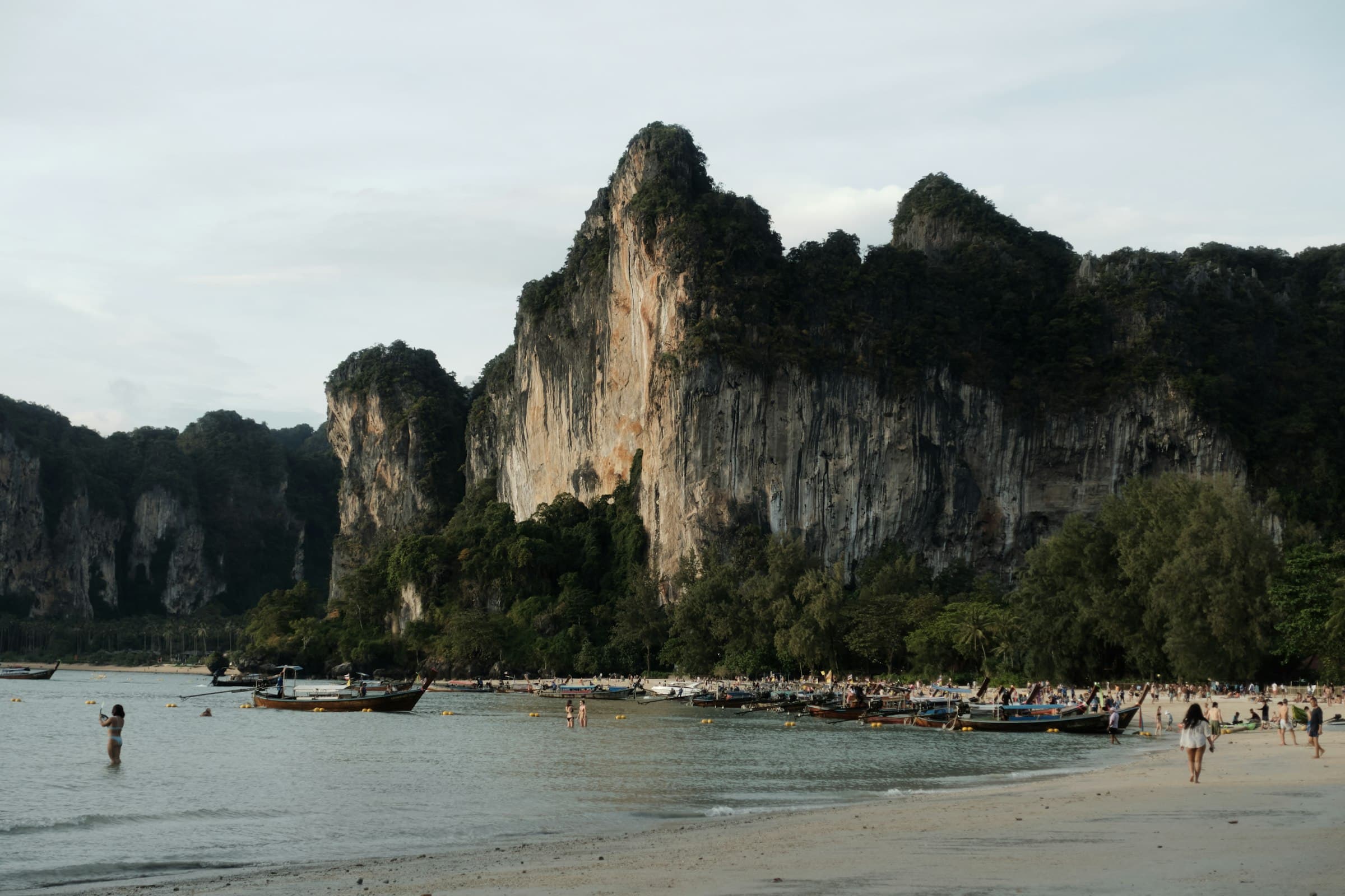 Longtail boats moored along a beach beneath a towering limestone cliff